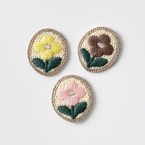 May include: Three embroidered flower patches in yellow, brown, and pink. Each oval patch features a flower with green leaves, set against a cream-colored background with a woven border. The patches are arranged on a white surface.