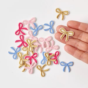 以下が含まれることがあります： ピンク、青、金など、さまざまな色の小さな装飾用リボンアップリケのコレクション。白い表面に散らばっており、手で持っているものもあります。リボンは布製で、ステッチされたアウトラインがあります。