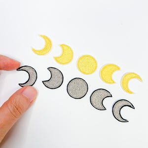 May include: Embroidered patches depicting the phases of the moon. The patches are in various shapes, including crescent moons and full moons, in shades of yellow and gray. The patches are arranged on a white background.