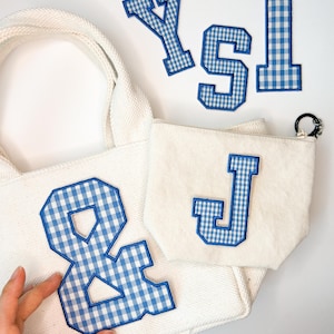 Può includere: Una borsa tote in tela color crema e una pochette con cerniera abbinata, entrambe caratterizzate da grandi toppe in stile varsity con motivo a quadretti blu e bianchi. Sono incluse ulteriori toppe sciolte nello stesso stile.
