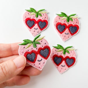 以下が含まれることがあります： 赤いハート型のサングラスをかけたピンクの刺繍されたイチゴのパッチが4つ。イチゴは緑の葉と笑顔をしています。