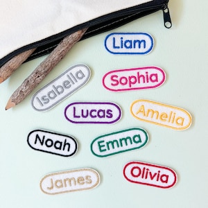 May include: Nine oval-shaped fabric name tags with embroidered names in various colors. The names are Liam, Sophia, Isabella, Lucas, Amelia, Noah, Emma, Olivia, and James.