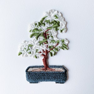 Könnte beinhalten: Gestickter Aufnäher eines Bonsai-Baums mit grünen Blättern und weißen Blüten in einem blauen Topf. Der Baum hat einen braunen Stamm und der Topf einen braunen Rand.
