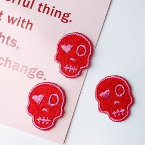 Könnte beinhalten: Drei rote gestickte Totenkopf-Patches mit rosa Herzen als Augen.