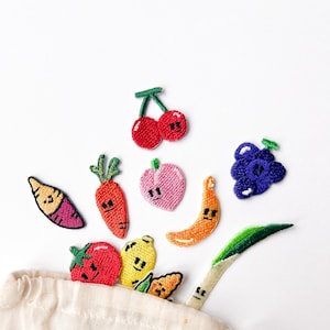 Puede incluir: Una bolsa blanca de cordón con el texto "Little Things Make" impreso en ella. La bolsa está llena de parches bordados de colores de varias frutas y verduras, incluyendo una cereza, un plátano, una zanahoria, una uva, un limón, una fresa, una batata y una cebolla verde.