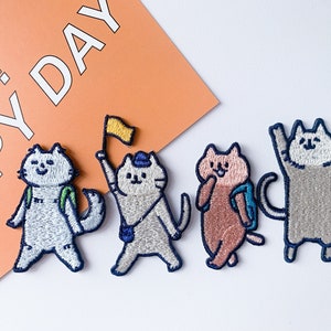 May include: Four embroidered iron-on patches featuring cartoon cats with backpacks. The cats are wearing different coloured clothing and have different expressions. The patches are perfect for adding a touch of whimsy to clothing, bags, or other fabric items.