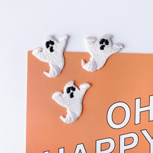 Könnte beinhalten: Drei weiße Filz-Geister-Applikationen mit schwarzen Augen auf einer pfirsichfarbenen Karte mit dem Text "OH! HAPPY" in Weiß.