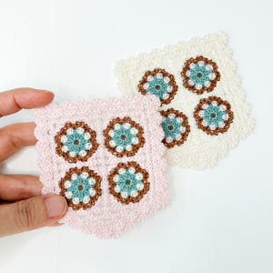 May include: Two handmade crochet coasters. One is pale pink, the other is off-white. Each coaster features four floral designs with teal, brown, and white accents. The edges of the coasters have a scalloped trim.