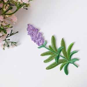 Può includere: Toppa ricamata di lavanda di colore viola e verde. Il fiore è dettagliato con più strati di petali e le foglie sono di un verde brillante.