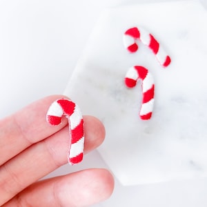 以下が含まれることがあります： 赤と白のストライプのミニチュアキャンディケーンが3つ。1つのキャンディケーンは人の手で持たれ、他の2つは白い表面にあります。キャンディケーンは、お祝いの休日の装飾です。