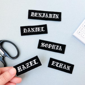 May include: Five black fabric patches with white embroidered lettering. The patches spell out the names "Benjamin", "Daniel", "Sophia", "Hazel", and "Ethan".
