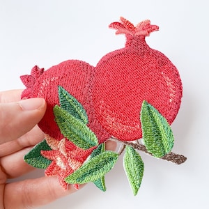 Puede incluir: Parche bordado de una granada roja con hojas verdes. La granada es un símbolo popular de abundancia y fertilidad.