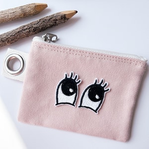 Könnte beinhalten: Eine rosa Reißverschlusstasche mit einer weißen Öse und zwei gestickten Augen mit schwarzen Wimpern.