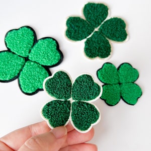 May include: Four green felt shamrock patches with white outlines. One shamrock is being held in a hand.