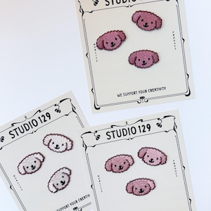 Può includere: Due carte con il testo "STUDIO 129" e la frase "WE SUPPORT YOUR CREATIVITY". Ogni carta presenta tre toppe ricamate termoadesive di cani barboncini rosa e bianchi.
