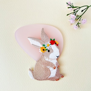 Könnte beinhalten: Ein braunes gesticktes Häschen mit weißem Gesicht und einer Blumenkrone. Die Krone hat rote, gelbe und weiße Blumen.