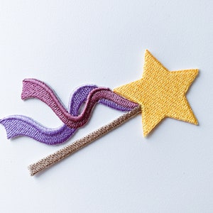 Puede incluir: Un parche bordado de una estrella dorada en un palo con serpentinas de cinta morada y rosa.