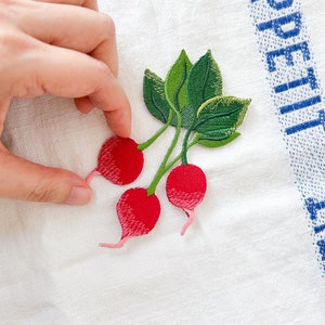 Puede incluir: Rábanos rojos bordados con hojas verdes sobre un fondo de tela blanca.