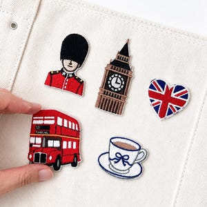 May include: A set of five embroidered patches featuring London-themed icons including a red double-decker bus, a Queen's Guard in a black bearskin hat, the Big Ben clock tower, a Union Jack heart, and a white teacup with a blue bow on a saucer.
