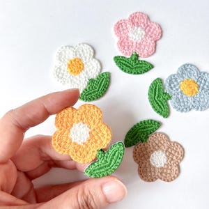 May include: A collection of colorful, crocheted flower appliques in shades of white, pink, blue, orange, and brown, each with a contrasting center. Green leaves accompany the flowers. The appliques are ideal for crafting and embellishment projects.