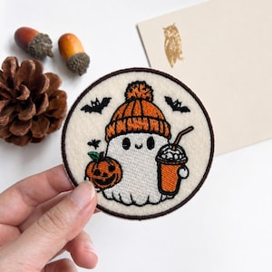 May include: A circular embroidered patch featuring a white ghost wearing an orange knit hat with a pom-pom. The ghost holds a small orange pumpkin and a cup with a straw. Three black bats are embroidered around the ghost on a cream background.