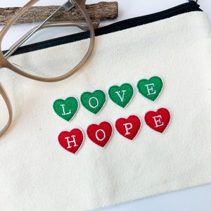 May include: A cream canvas pouch with a black zip, embroidered with green hearts spelling "LOVE" and red hearts spelling "HOPE." A pair of spectacles with a brown frame and a small wooden stick are placed on top.