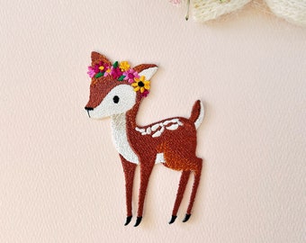 以下が含まれることがあります： 刺繍されたアイロン接着パッチ。茶色の鹿が花冠をかぶっています。鹿は4本足で立っており、頭は左を向いています。花冠はピンク、黄色、白の花でできています。