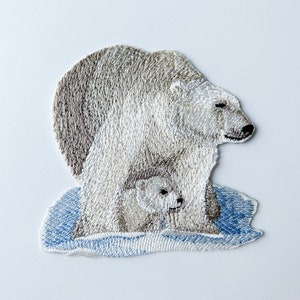May include: Embroidered patch of a polar bear and cub standing on a blue ice floe. The bear is white with brown fur on its back and the cub is white.