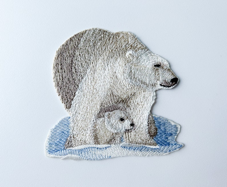 May include: Embroidered patch of a polar bear and cub standing on a blue ice floe. The bear is white with brown fur on its back and the cub is white.