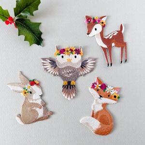 May include: Embroidered patches featuring woodland animals: a deer, owl, fox, and rabbit, each adorned with a floral crown. The patches are in shades of brown, white, and orange, with green holly leaves in the upper left corner.