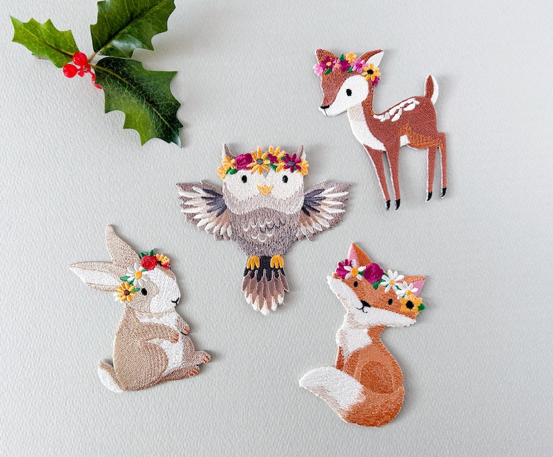 May include: Embroidered patches featuring woodland animals: a deer, owl, fox, and rabbit, each adorned with a floral crown. The patches are in shades of brown, white, and orange, with green holly leaves in the upper left corner.