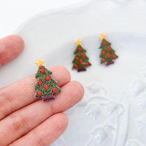 Peut inclure: Trois petites décorations de sapin de Noël brodées en feutre vert, avec des boules en feutre rouge et des étoiles en feutre jaune. Les décorations sont sur une surface blanche.