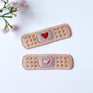 May include: Two embroidered iron-on patches shaped like bandages with a heart in the center. One patch has a red heart and the other has a pink heart.