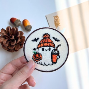 May include: A circular embroidered patch with a white ghost wearing an orange beanie, holding a pumpkin and a drink. The patch has a brown border and three bats. A fun, seasonal accessory for autumn.