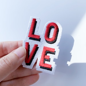 Op de afbeelding: Een witte stoffen patch met het woord "LOVE" geborduurd in rode en zwarte draad. De letters zijn zwart omrand en rood gevuld.