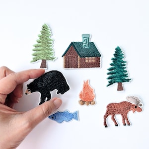 以下が含まれることがあります： 黒い熊、緑の屋根の小屋、キャンプファイヤー、ヘラジカ、青い魚、2本の常緑樹が刺繍されたパッチ。パッチはさまざまな色で、白い背景に配置されています。