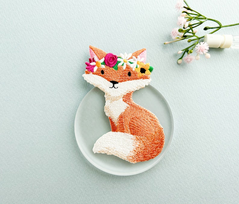 May include: Embroidered patch of a sitting fox with orange and white fur, wearing a floral crown with pink, yellow, and white flowers. The fox is on a clear, round surface against a light blue background.
