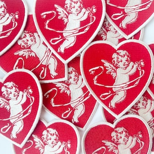 May include: A collection of red heart-shaped patches featuring white embroidered cherubs holding ribbons. The cherubs are depicted in a vintage style.