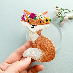 May include: Embroidered patch of a seated fox with orange and white fur, wearing a floral crown of pink, yellow, and white flowers. The fox has a black nose and eyes. The patch is likely for clothing or accessories.