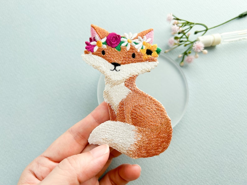 May include: Embroidered patch of a seated fox with orange and white fur, wearing a floral crown of pink, yellow, and white flowers. The fox has a black nose and eyes. The patch is likely for clothing or accessories.