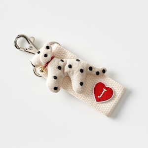 May include: A handmade keychain featuring a plush Dalmatian dog with black spots, a red collar, and a small gold bell. The dog rests on a cream-colored fabric with a red heart patch and the letter "J".