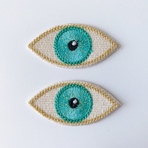 May include: Two embroidered eye patches with a teal blue eye and a black pupil. The eye patches are on a white background with a gold outline.