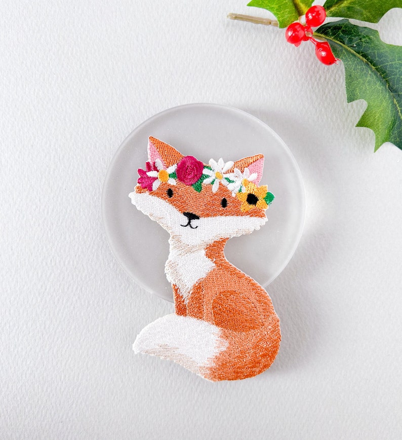 May include: Embroidered patch featuring a cartoon fox with orange fur, white chest, and a floral crown. The fox is sitting on a clear, round surface. The patch has a detailed design with various colors, including pink, yellow, and green.