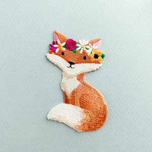 May include: Embroidered patch of a fox with orange and white fur, wearing a floral crown with pink, white, and yellow flowers. The fox is sitting with its tail curled. The patch is on a light blue background.