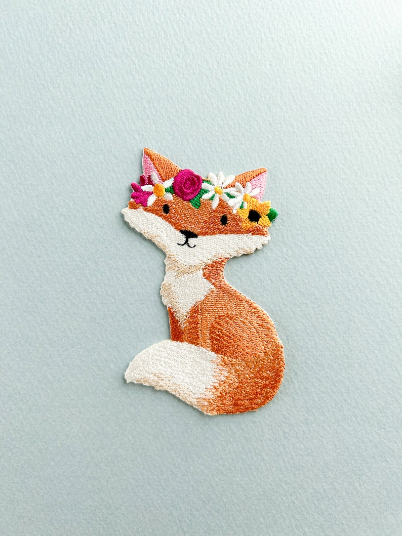 May include: Embroidered patch of a fox with orange and white fur, wearing a floral crown with pink, white, and yellow flowers. The fox is sitting with its tail curled. The patch is on a light blue background.