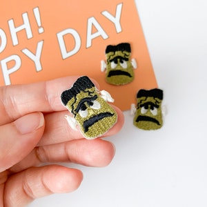 May include: Three green and black embroidered iron-on patches featuring a cartoon monster with a grumpy expression. The patches have white stitching around the eyes and mouth.