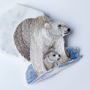 May include: Embroidered patch of a mother polar bear and her cub standing on a blue ice floe. The polar bears are white with brown fur details.