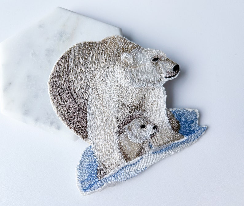May include: Embroidered patch of a mother polar bear and her cub standing on a blue ice floe. The polar bears are white with brown fur details.