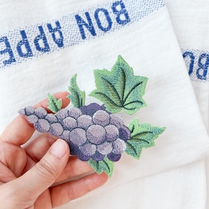 Può includere: Primo piano di un grappolo d'uva ricamato viola con foglie verdi. Il ricamo è su un tessuto bianco a strisce blu e bianche con il testo "BON APPETIT" in blu.