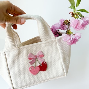 May include: A small, white canvas tote bag with a pink bow and two cherries embroidered on the front. The cherries are red and pink.